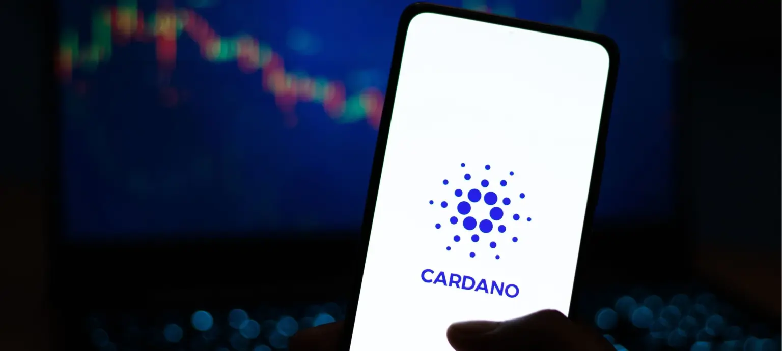 Hand holding a smartphone displaying the Cardano logo with a crypto price chart in the background
