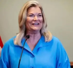 Woman in a blue blouse smiling while speaking at a podium microphone with an American flag beside her