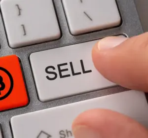 Finger pressing a computer keyboard sell key beside a red button displaying the Bitcoin symbol