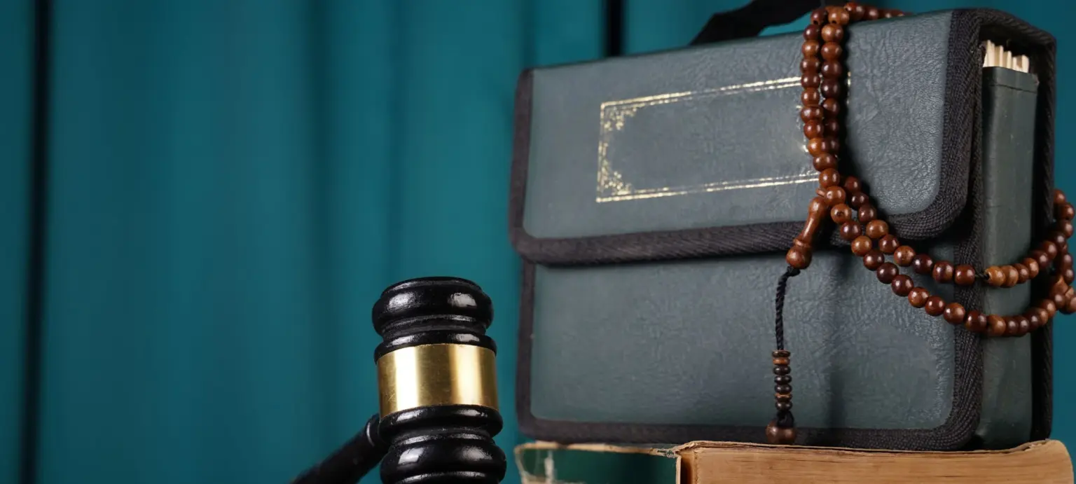 Judge’s gavel beside Islamic book and beads against a teal background.