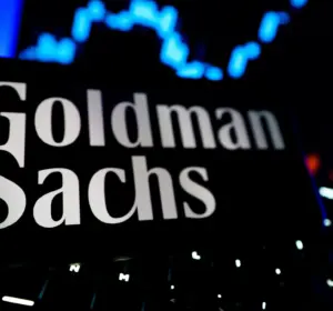 Goldman Sachs name displayed on a screen with a blue financial market chart in the background.