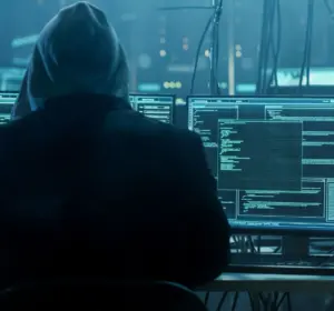 Hooded figure viewed from behind sitting at multiple monitors showing code and a world map