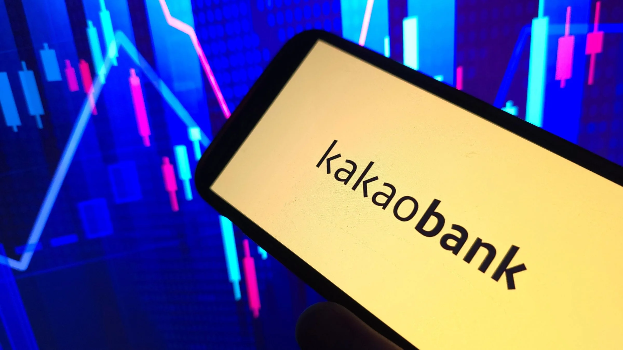 Smartphone displaying the KakaoBank name in front of colorful market chart graphics on a dark background.