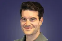 Studio headshot of a smiling man with dark, wavy hair, wearing a gray sweater, against a deep purple-blue gradient background.