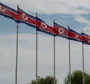 Row of North Korean flags waving on tall flagpoles against a cloudy blue sky