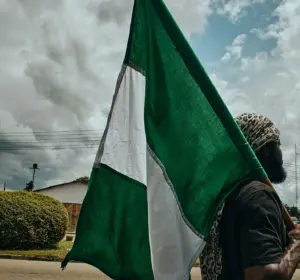 Person carrying a Nigerian flag along a roadside under heavy clouds with a statue visible in the background.