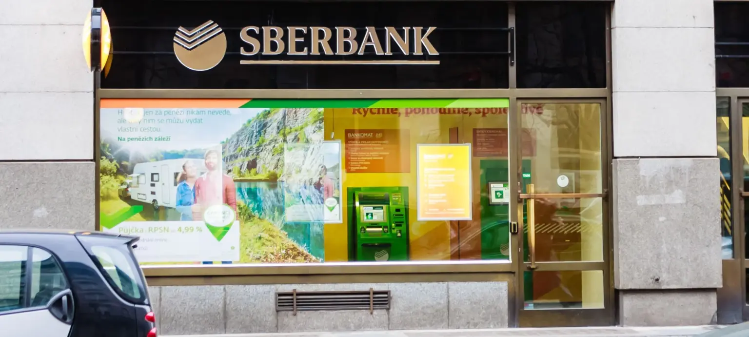 Sberbank branch exterior with illuminated logo, glass entrance, advertising displays, and an ATM beside the doorway.