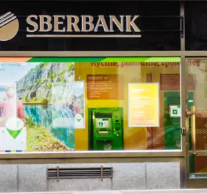Sberbank branch exterior with illuminated logo, glass entrance, advertising displays, and an ATM beside the doorway.