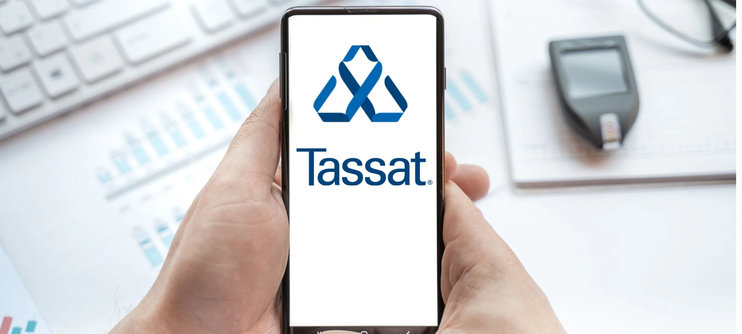 Hands holding a smartphone displaying the Tassat logo above financial charts