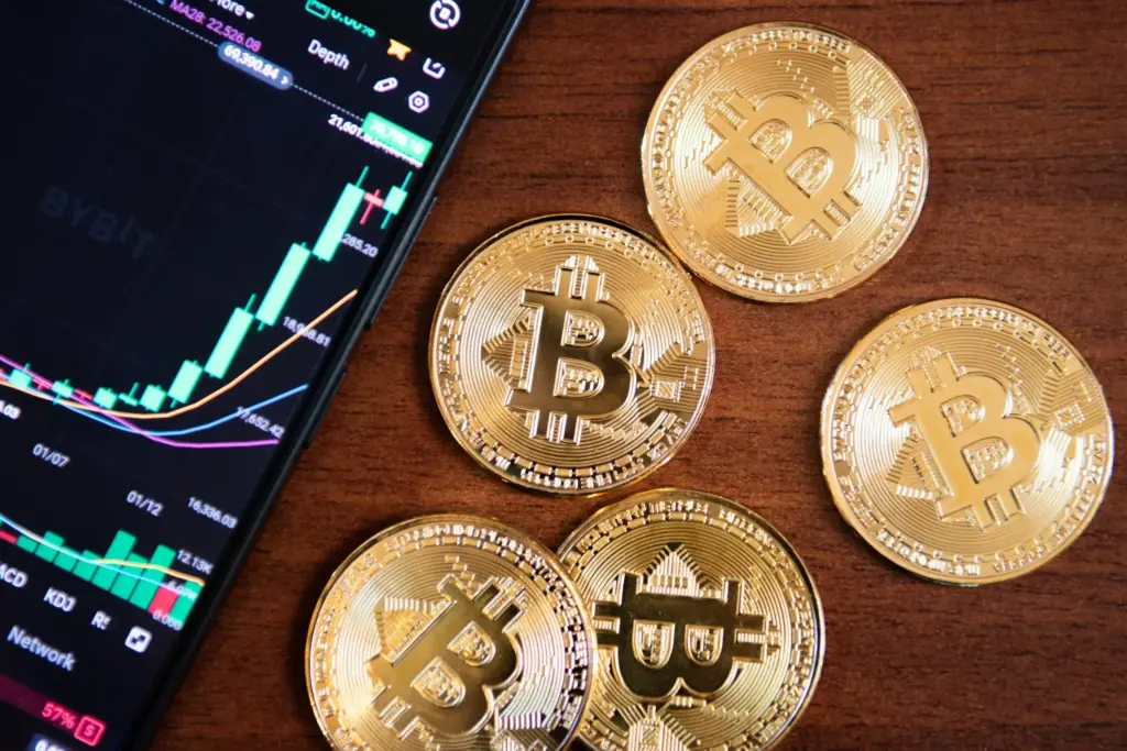 Gold Bitcoin tokens beside a smartphone displaying an upward cryptocurrency trading chart on a wooden surface