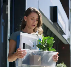 Woman holding a plastic box with office items and a potted plant outside a modern building.