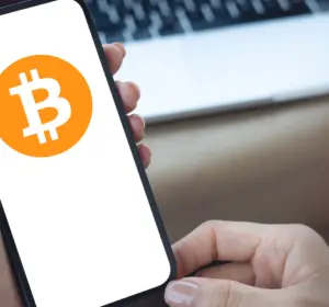 Person holding a smartphone displaying the Bitcoin logo, with a laptop in the background